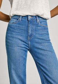 Jean taille haute bleue en denim, dotée de cinq poches, fermeture à bouton en métal et couture contrastée. Texture lisse avec une légère élasticité.