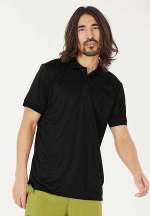 Homme aux cheveux mi-longs portant un polo noir et un pantalon vert olive, debout devant un fond clair uni.