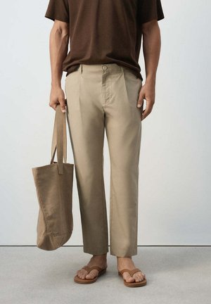 Persona con pantalones beige y sandalias marrones sosteniendo con una mano un bolso tote beige a juego contra un fondo liso.