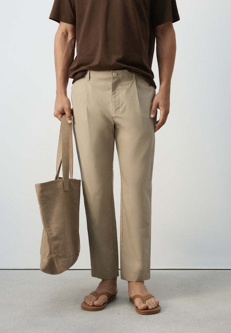 Personne portant un pantalon beige et des sandales marron tenant un sac cabas beige assorti d'une main sur un fond uni.