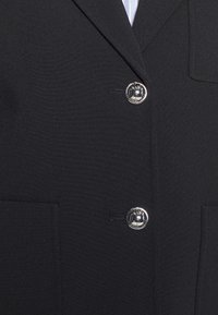 Blazer noir sur mesure avec deux boutons argentés brillants et une petite poche poitrine visible sur le côté droit.