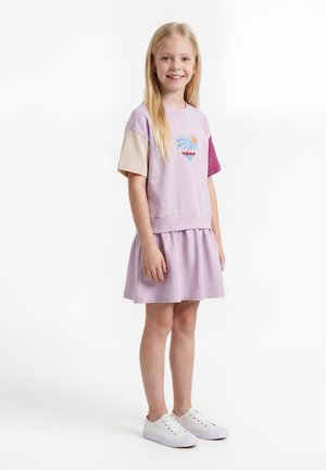 Lachend meisje met lang blond haar draagt een pastelpaars jurkje met meerkleurige mouwen en witte sneakers, staand tegen een witte achtergrond.
