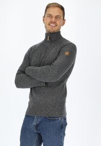 Maglione a coste grigio con collo alto, dotato di chiusura con zip e un piccolo emblemma sulla manica sinistra. Indossato con jeans blu.