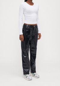 Person i vitt långärmad crop-top, svarta imitation läder-texturerade byxor och vita sneakers med gröna detaljer, som står mot en neutral bakgrund.