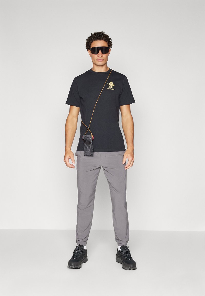 Schwarzes T-Shirt mit einem goldenen Grafikdesign, graue tapered Hosen und schwarze Sneaker. Accessoires umfassen übergroße Sonnenbrillen und eine kleine Umhängetasche an einem Band.
