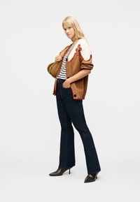 Ξανθιά γυναίκα που φορά καφέ και κρεμ ζακέτα, ριγέ πουκάμισο, σκούρα φαρδιά τζιν και μαύρα μπότες με τακούνι, κρατώντας μια ταμπά τσάντα με υφή.