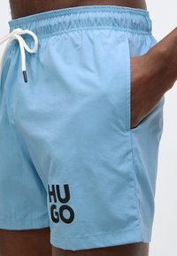 Világoskék fürdőnadrág, amely rugalmas derékpánttal, húzózsinóros záródással és egy fekete "HUGO" logóval rendelkezik a bal alsó oldalon.