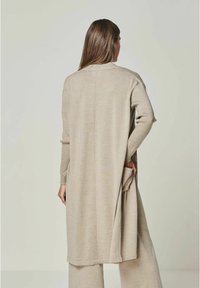 Beige langer Cardigan mit lockerer Passform, ausgestattet mit Seitentaschen und einer geraden Rückennäht, aus einem weichen, strukturierten Stoff gefertigt.
