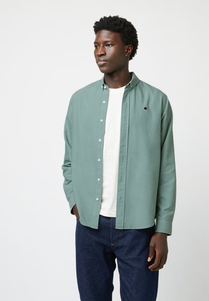 Groene button-up shirt met een zachte textuur en een klein geborduurd ontwerp, gedragen over een wit T-shirt en donkere jeans. Casual pasvorm.