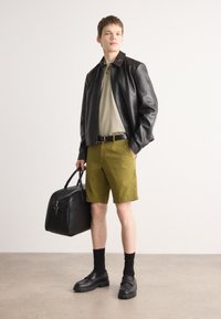 Svart lærjakke, olivengrønne shorts, svart belte, slip-on sko og svarte sokker. Holder en svart duffelbærepose. Enkel bakgrunn.