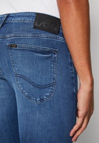Jeans de denim azul con una textura suave, que cuentan con un parche de logo de cuero negro y un único bolsillo trasero con un detalle de costura curva.