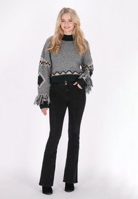 Grauer gestrickter Pullover mit gemusterten Ärmeln, fransigen Bündchen und einem grünen Band. Kombiniert mit schwarzen ausgestellten Jeans und schwarzen High Heels.