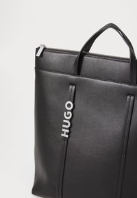 Borsa tote in pelle nera con due manici superiori, chiusura con zip, dettagli con logo in rilievo e superficie liscia. Presenta un design minimalista e un aspetto elegante.