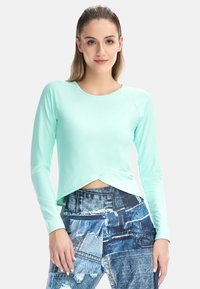 Ženska s ravnimi lasmi, oblečena v svetlo modro majico z dolgimi rokavi in kavbojke iz različnih delov džinsa, stoji ob belem ozadju.