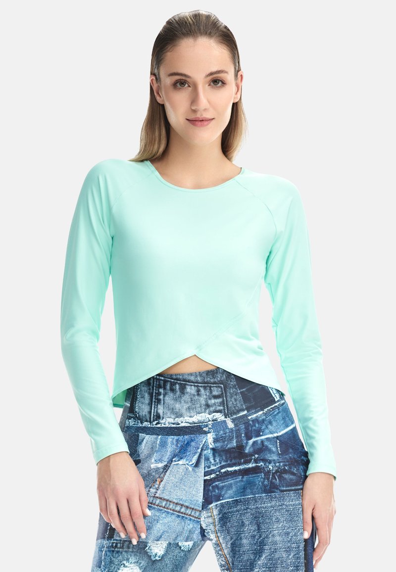 Ženska s ravnimi lasmi, oblečena v svetlo modro majico z dolgimi rokavi in kavbojke iz različnih delov džinsa, stoji ob belem ozadju.