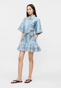 Νεαρή γυναίκα που φοράει ένα κοντό, ανοιχτό μπλε φόρεμα με σχέδια και δεμένη μέση, στέκεται με τα χέρια στις τσέπες της και μαύρα σαγιονάρες, πάνω σε έναν απλό φόντο.