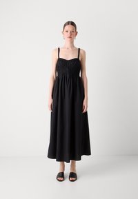 Robe maxi noire avec bretelles fines ajustables, corsage froncé et jupe fluide. Tissu en coton lisse, longueur cheville, associée à des sandales à glissière noires.