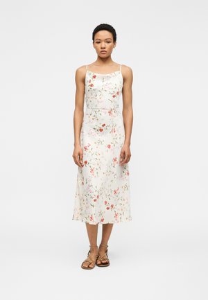 Sieviete stāv, valkājot balvu midi ziedu kleitu ar plānām lencēm un pieskaņotu jostas siksnu viduklī, kombinēta ar brūnām sandaletēm ar siksnām.