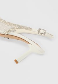 Elfenbensfarvet sandal med høje hæle med en gennemsigtig overdel pyntet med rhinsten, en firkantet spænde og en slank hæl med en beige spids.