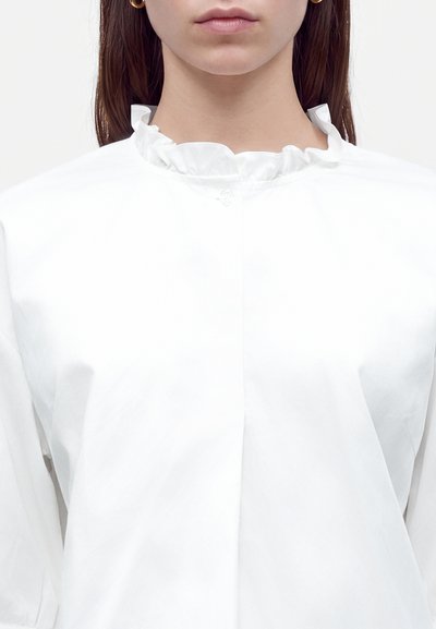 Femme portant une blouse blanche à col volanté et des boucles d'oreilles créoles dorées, montrée des épaules aux lèvres sur un fond uni.