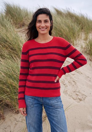 Mujer sonriente está de pie en una playa de arena con hierba alta, vistiendo un suéter rojo con rayas oscuras y vaqueros azules, con la mano en la cadera.