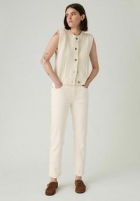Kerma neuleliivi, jossa on teksturoituja kuvioita ja puunappulat, yhdistettynä kermaisiin slim-fit-housuihin ja ruskeisiin mokkasiinikenkiin.