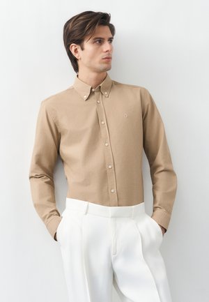 Hombre vistiendo una camisa beige abotonada y pantalones blancos plisados, de pie con las manos en los bolsillos, mirando hacia un lado contra un fondo blanco.