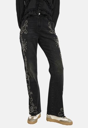 Vaqueros negros de pierna recta con estampados florales y espirales con tachuelas plateadas, llevados con zapatillas beige con detalles de purpurina y una camiseta negra con volantes.