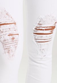 Jeans blancos desgarrados que presentan dos parches prominentes en las rodillas con bordes deshilachados y un acabado texturizado.