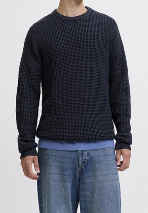 Homme portant un pull en maille gaufrée bleu foncé par-dessus une chemise bleu clair avec un jean bleu ample, debout devant un fond blanc.