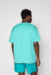 Camiseta de algodón de color turquesa claro con mangas cortas, escote redondo y un corte relajado. La parte posterior es lisa, sin logotipos ni patrones visibles.