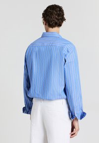 Uomo con capelli ricci castani che indossa una camicia a maniche lunghe blu a righe e pantaloni bianchi a trama, di spalle su uno sfondo grigio uniforme.