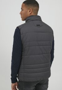 Vest gri cu umplutură, cu guler înalt, având sub el un pulover texturat de culoare bleumarin. Modelul matlasat este vizibil, iar pe spate se află o emblema.