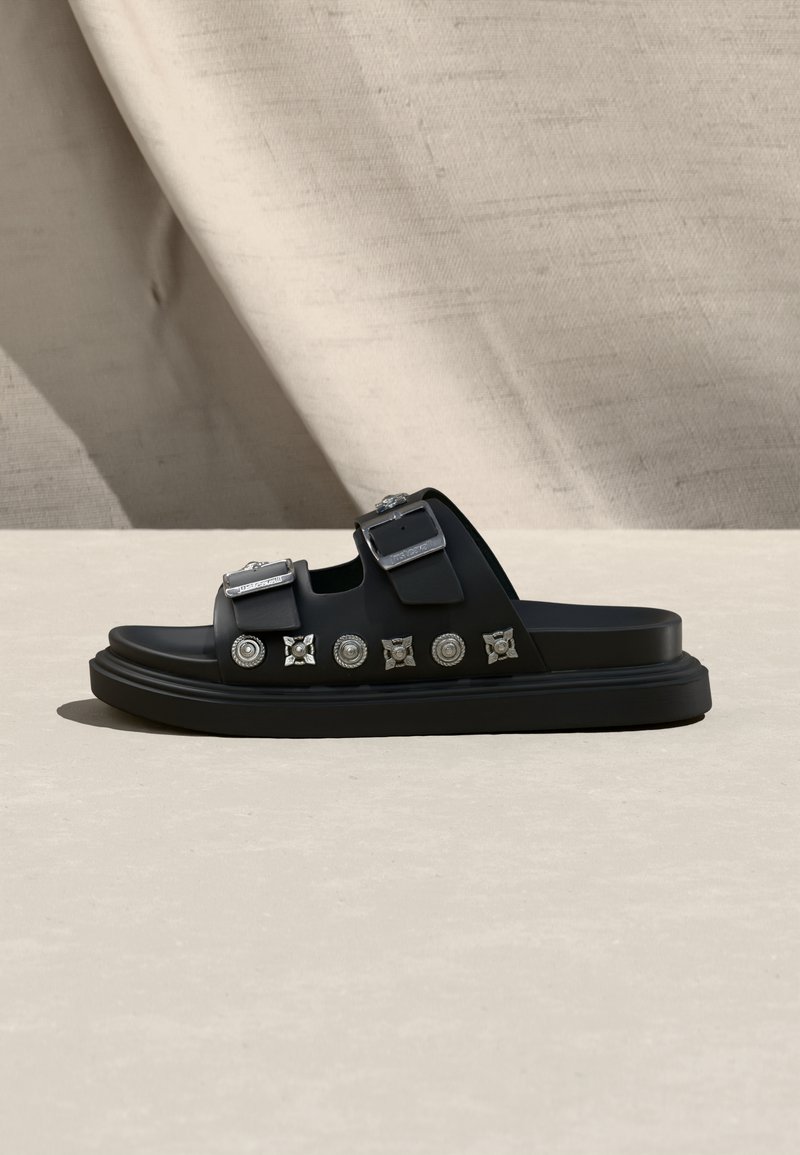 Μαύρο πλατφόρμα σανδάλι με δύο ρυθμιζόμενες ασημένιες αγκράφες και διακοσμητικά ασημένια καρφιά στο πλάι, τοποθετημένο πάνω σε ουδέτερο υφασμάτινο φόντο.