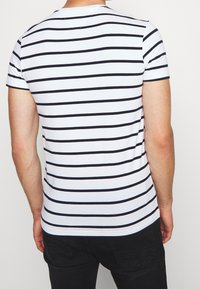 Mężczyzna w białej koszuli z krótkim rękawem w poziome czarne paski, widoczny z tyłu, w połączeniu z czarnymi spodniami.