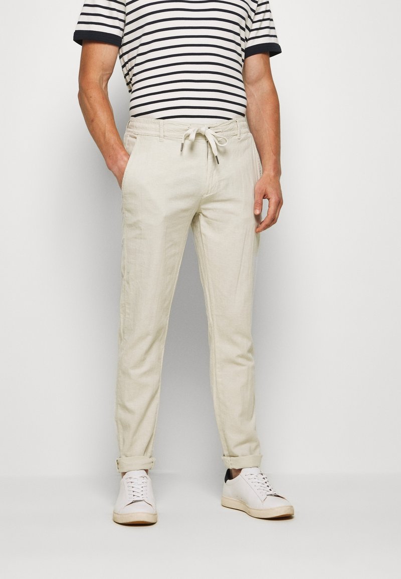 Pantalon beige en mélange de coton et lin, coupe fuselée, taille élastique avec cordon de serrage, et revers roulés, associé à une chemise à manches courtes rayée et des baskets blanches.