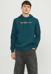 Sudadera color teal con un bolsillo tipo canguro en la parte delantera, capucha con cordón y un diseño gráfico frontal en naranja y blanco que dice "JACK & JONES". Tejido de mezcla de algodón.