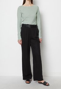 Frau trägt ein grün-weiß gestreiftes Langarmoberteil, hochgeschnittene schwarze weite Hosen mit einem schwarzen Gürtel und schwarze Slide-Sandalen.
