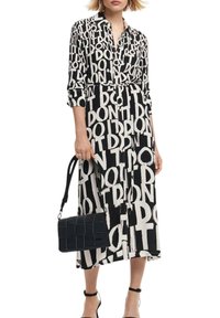 Abito lungo in bianco e nero con un audace motivo testuale, colletto, chiusura con bottoni sul davanti e vita con cintura; abbinato a una borsa a tracolla nera testurizzata.