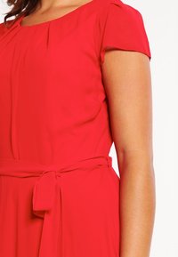 Robe rouge à manches courtes avec un col rond et une ceinture à la taille. Tissu lisse avec un léger éclat, présentant des plis au niveau du corsage.