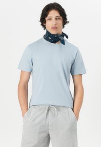 T-shirt en coton bleu clair avec un col rond et des manches courtes, arborant un petit logo sur la poitrine. Associée à un short rayé.