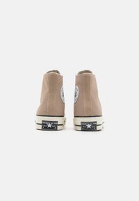 Baskets montantes en toile couleur marron clair, avec une semelle en caoutchouc, un logo classique noir et blanc, et un patch logo circulaire de chaque côté.