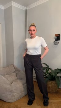 Witte T-shirt met korte mouwen, donkere broek met hoge taille en zwarte schoenen. De achtergrond bestaat uit een lichtgrijze muur, een zachte stoel en een potplant.