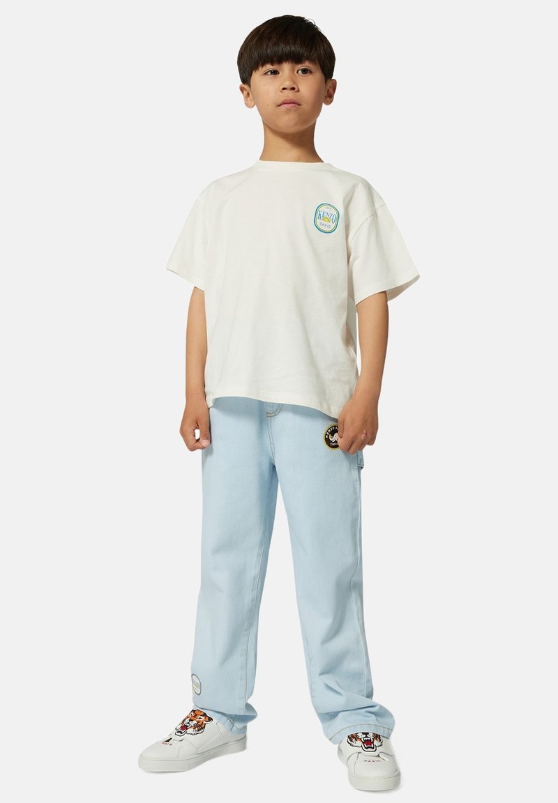 T-shirt en coton blanc avec un logo circulaire, pantalon en jean bleu clair et baskets blanches avec des graphismes de tigre brodés.