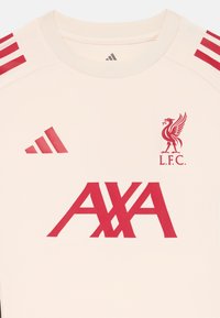 Krämfärgad tr�öja med röda horisontella remsor på axlarna, Liverpool FC-logotyp och "AXA" tryckt framträdande i rött.