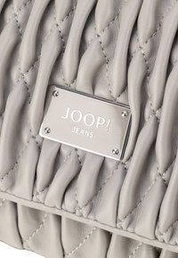 Siva usnjena torbica s pikčasto teksturo, ki ima osrednjo srebrno logotipno ploščico z napisom "JOOP! JEANS." Vidne so šivane podrobnosti.