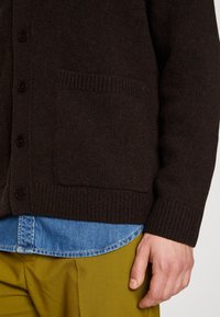 Brązowy wełniany kardigan z guzikami, ściągaczami przy rękawach i kieszenią, nałożony na niebieską koszulę dżinsową i żółte spodnie.