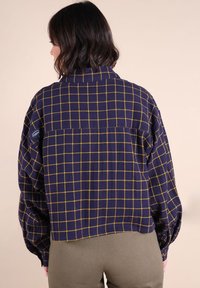 Femme de dos portant une veste courte à carreaux bleu marine avec des lignes jaunes et un pantalon vert olive sur un fond clair.