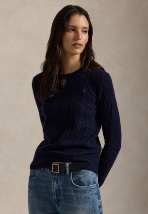 Nő tengerészkék, kötött mintás pulóverben és kék farmernadrágban, fekete övvel, egy egyszínű bézs háttér előtt áll.