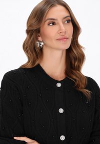 Cardigan nero a maglia grossa con motivo testurizzato e decorazioni in perle nere. Chiusura frontale con bottoni e dettagli in tonalità argento.